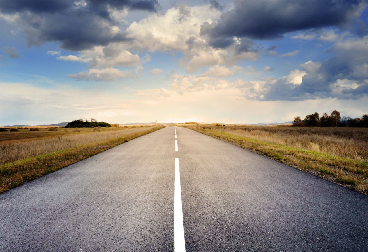 A long straight road stretches through fields under a dramatic cloudy sky.